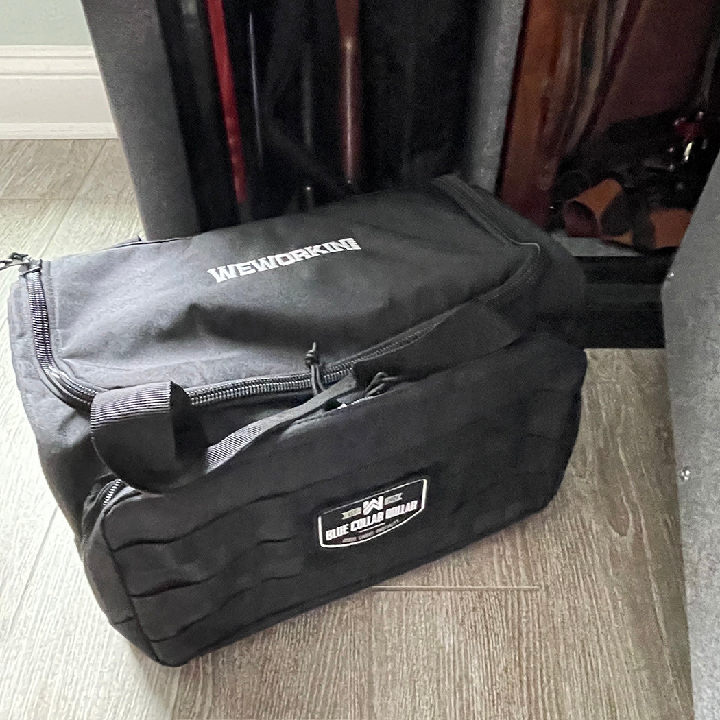 Stealth Black tactical range/gear bag pictured from front beside an open gun safe. WEWORKIN BRAND logo embroidered in white thread on the top panel. 600D polyester canvas, Zippered D-shaped main compartment, Front zippered pocket with daisy chain/loop panel for patches (Blue Collar Dollar patch shown on range bag, all patches sold separately).