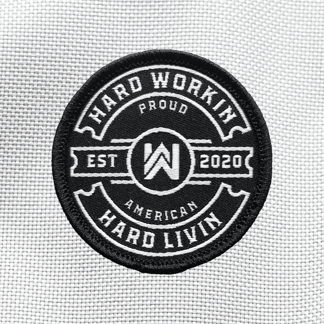 "HARD WORKIN. HARD LIVIN." text circling the WW icon logo and "EST 2020"  text in center, on a velcro-backed patch (both the hook and loop velcro sides included). [1] thread color for the design (white) on a black woven background, with black merrowed border. "PROUD AMERICAN" circles top and bottom under outer text. 2.25" wide circular Woven patch displayed on a grey canvas background.