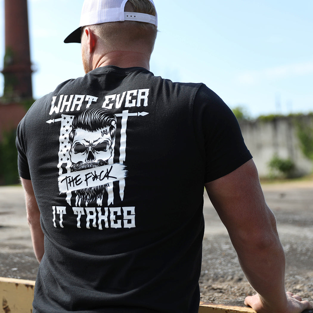 Man pictured from back wearing a We Workin graphic tee in Dark Black color. Short sleeve shirt imprinted with a WeWorkin designed graphic—the text "What Ever the F*CK It Takes" above and below a bearded skull overlaid on top of a tattered US Flag, hanging vertically on a rod (printed in white ink.) He is also wearing the WeWorkin Black/White Retro Trucker hat. 