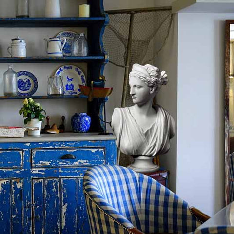 Photo of plaster cast sculpture of a bust of the goddess Diana with a checkered blue chair in the foreground and a weathered blue shelf with dishes and jugs to the left