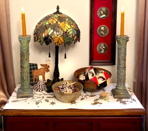 photo of small table with patterned, white tablecloth, two verdigris-color plaster candlesticks with lit orange candles, colorful glass lamp, decorative bowls with pine cones and a Venetian mask, pillow with moose image, and on off-white wall behind, a tall, narrow shadow box with three silver portrait medallions on red background