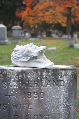 photo of plaster cast sculpture of a satyr head atop a gray headstone in the foreground in a cemetery