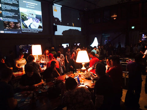 Games being played on tables lit by lamps while bystanders watch over on ShowDown Game Night at the Folsom Street Foundry.