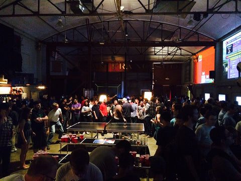 Crowd gathers around the table tennis game in the low light at the Folsom Street Foundry Game Night. 