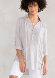 [Color: Grey/Ivory] A model wearing a relaxed yarn dye popover tunic shirt in a grey and ivory stripe. With long rolled sleeves, a button front, and two patch pocket details. 