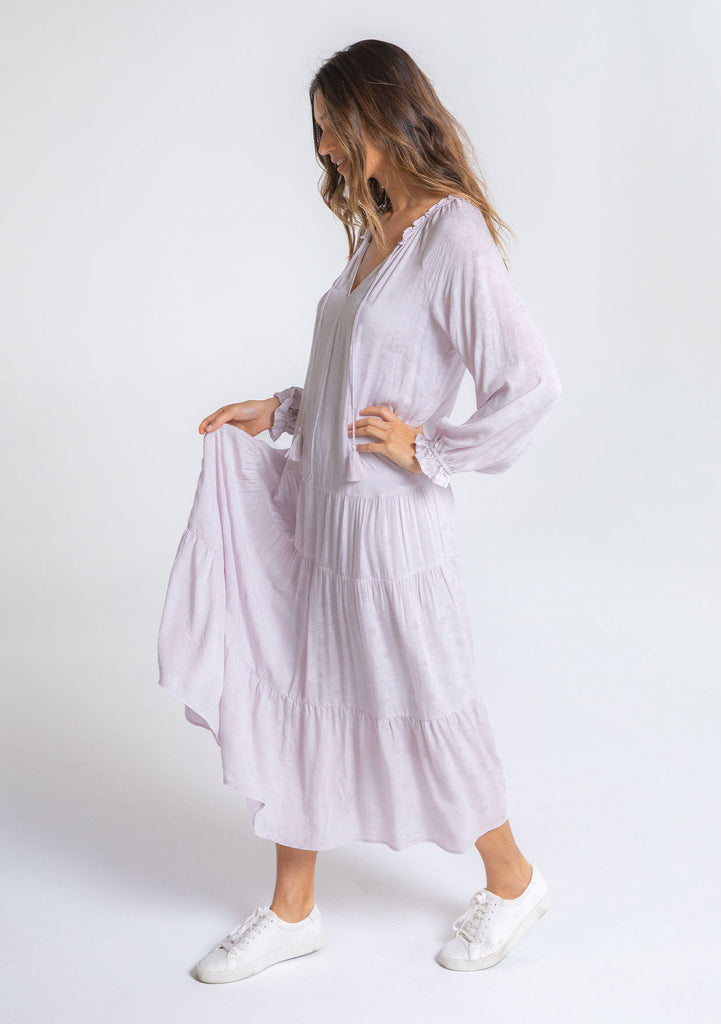 [Color: Orchid Tint] A model wearing an elegant and classic light purple bohemian peasant maxi dress in floral jacquard. Featuring a dropped waist, a tiered skirt, and a split v neckline with tassel ties.