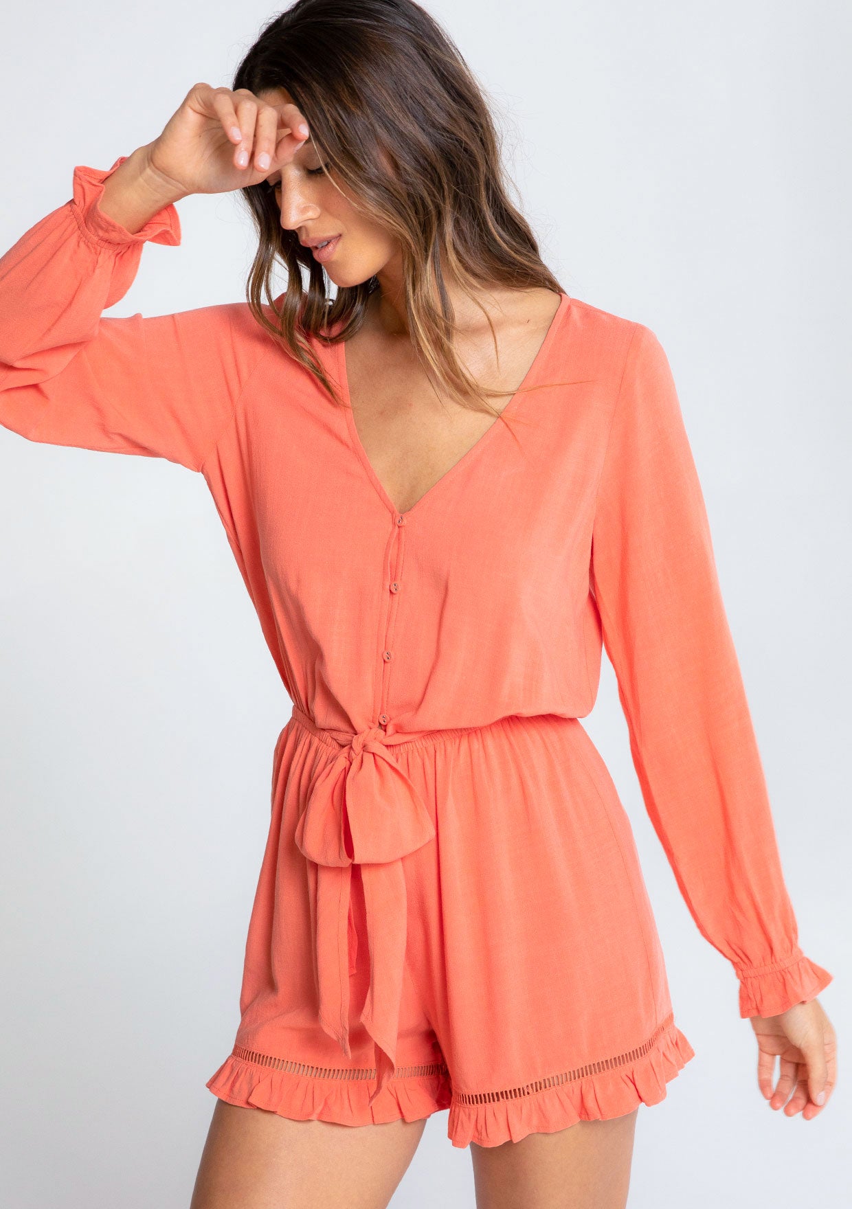 [Color: Papaya] A model wearing a coral ruffled long sleeve short romper made from a linen blend. With a tie front waist detail. 