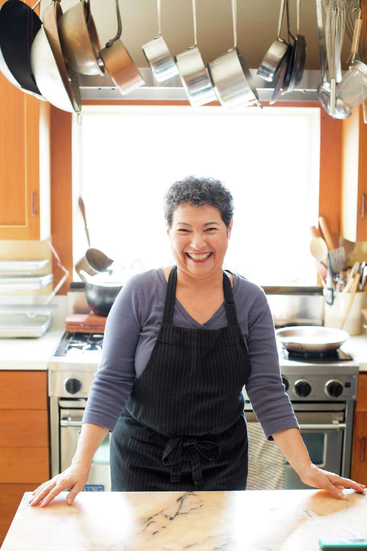 A woman with close cropped, greying hair smiles brightly, while standing in her kitchen.