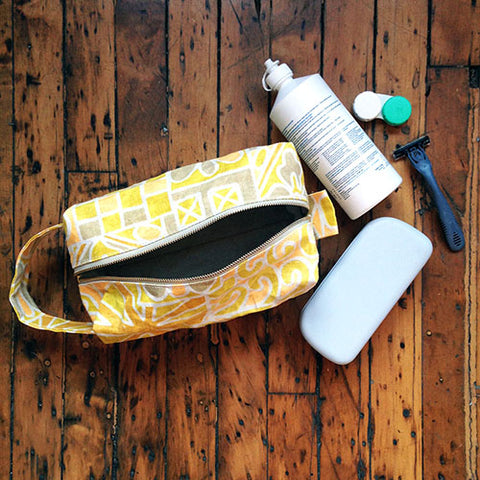 yellow patterned toiletry bag with contact solution bottle, contact case, and glasses case next to them on a wood floor