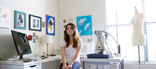 Photo of Jenny Nemlekar sitting at her computer desk with smaller sewing machine and mannequin behind her