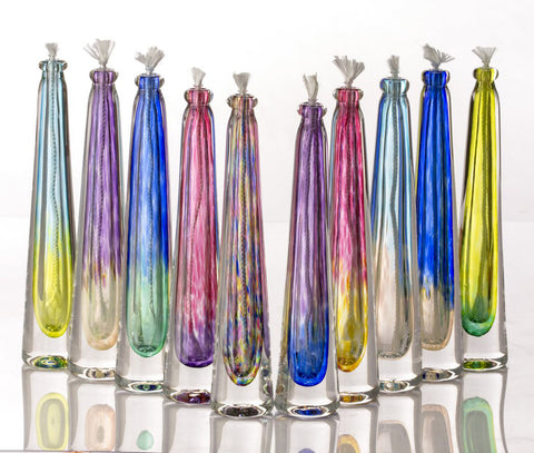 Eight tapered hand blown glass candles with ashes in a row with cotton wicks in a rainbow of colors.