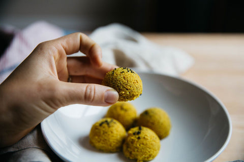 A hand holds one golden mylk macaroon with black lava salt sprinkled on top. Four more macaroons site on a white plate in the background.