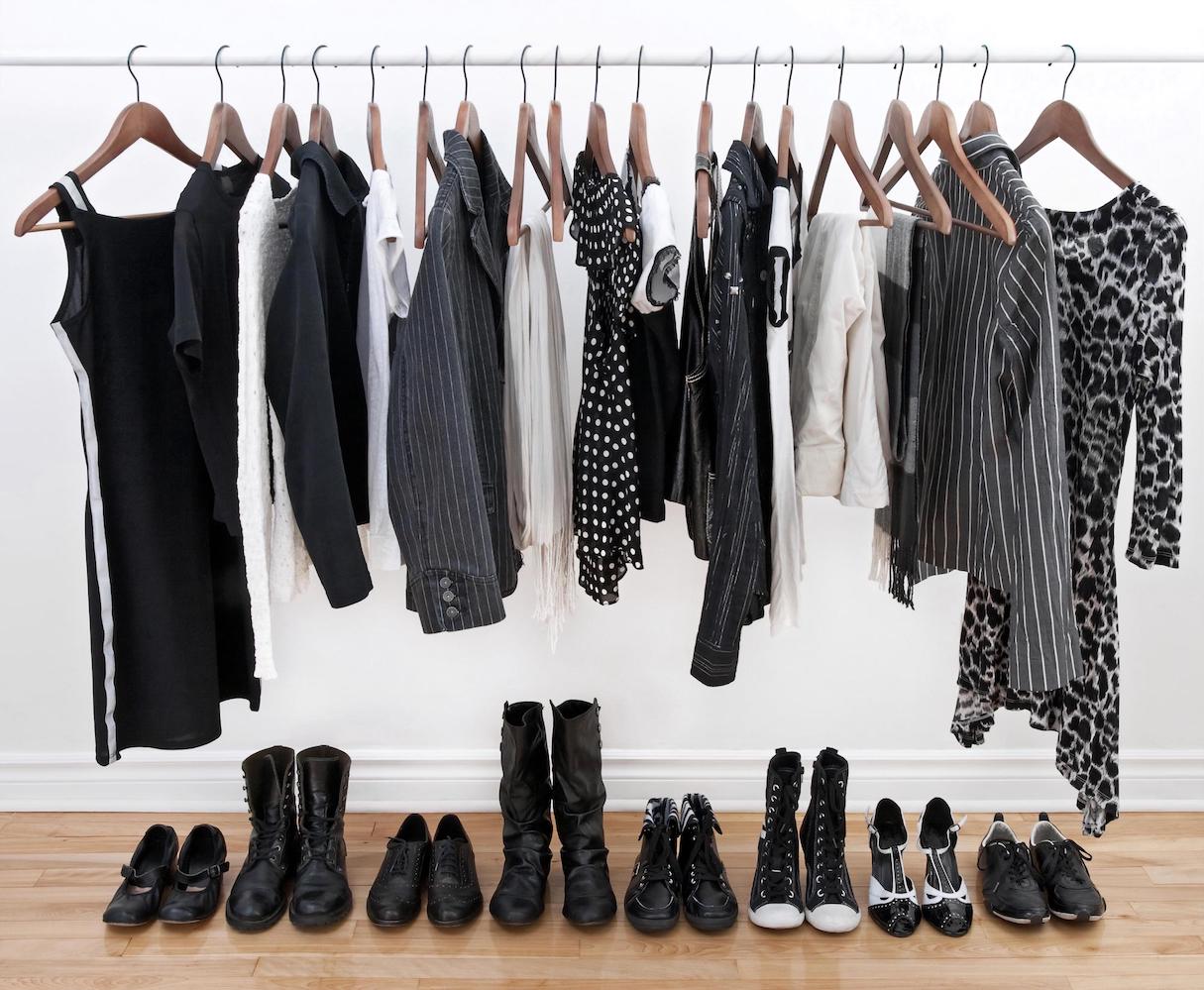 An assortment of black and white clothes hanging on a rack. Black and white shoes neatly lined up underneath the rack.
