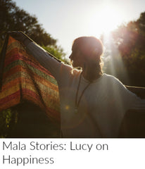 A woman with curly light brown hair holds out a shawl backlit by the sun. She wears a calcite moonstone and sandalwood mala necklace around her neck from Mama Malas