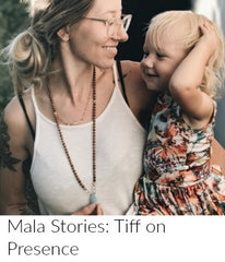 A young woman with tattoos and clear glasses holds her young blonde daughter in one arm, and holds the Amazonite guru stone of her sandalwood mala bead necklace in the other hand.