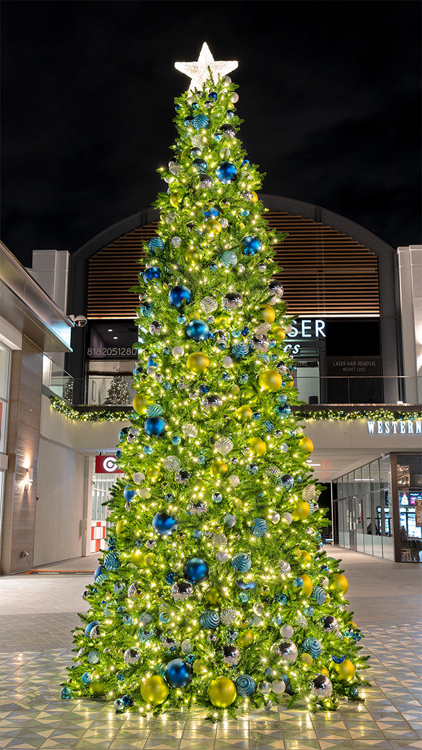 A brightly lit Christmas tree stands in the center of an outdoor shopping center. The tree is decorated with blue, green, and silver ornaments and a star on top.