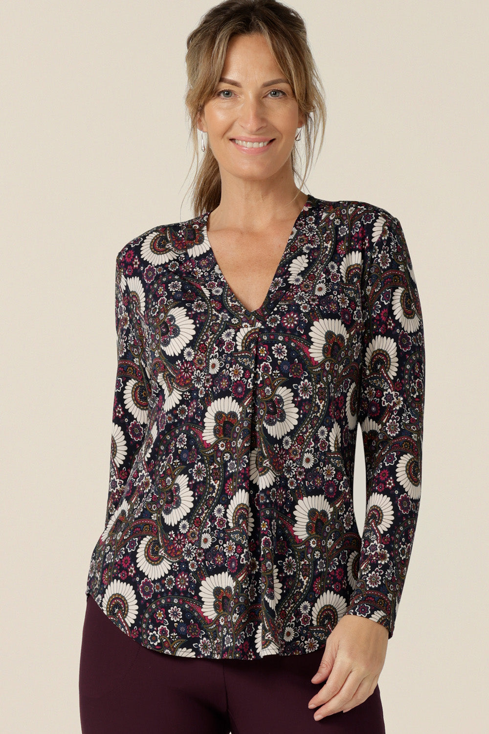 A size 10 woman wears a long-sleeve, V-neck top in paisley print jersey. Made in Australia by Australian and New Zealand women's clothing label, L&F this tailored work top is available to shop in sizes 8 to 24.