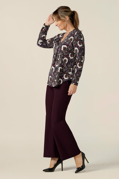 A size 10 woman wears a long-sleeve, V-neck top in paisley print jersey. Worn with flared leg, tailored trousers in Mulberry, this comfortable work top is made in Australia by Australian and New Zealand women's clothing label, L&F.