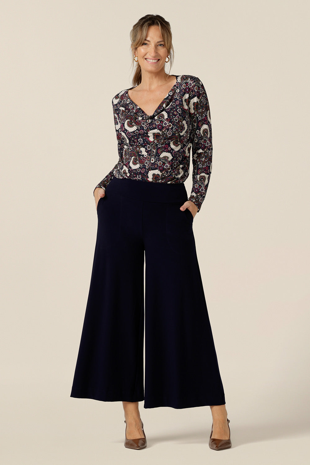 A size 10 woman wears a long sleeve women's cowl neck top in paisley print jersey. Worn with wide-leg navy pants, this top is a comfortable top for work and weekend wear. 