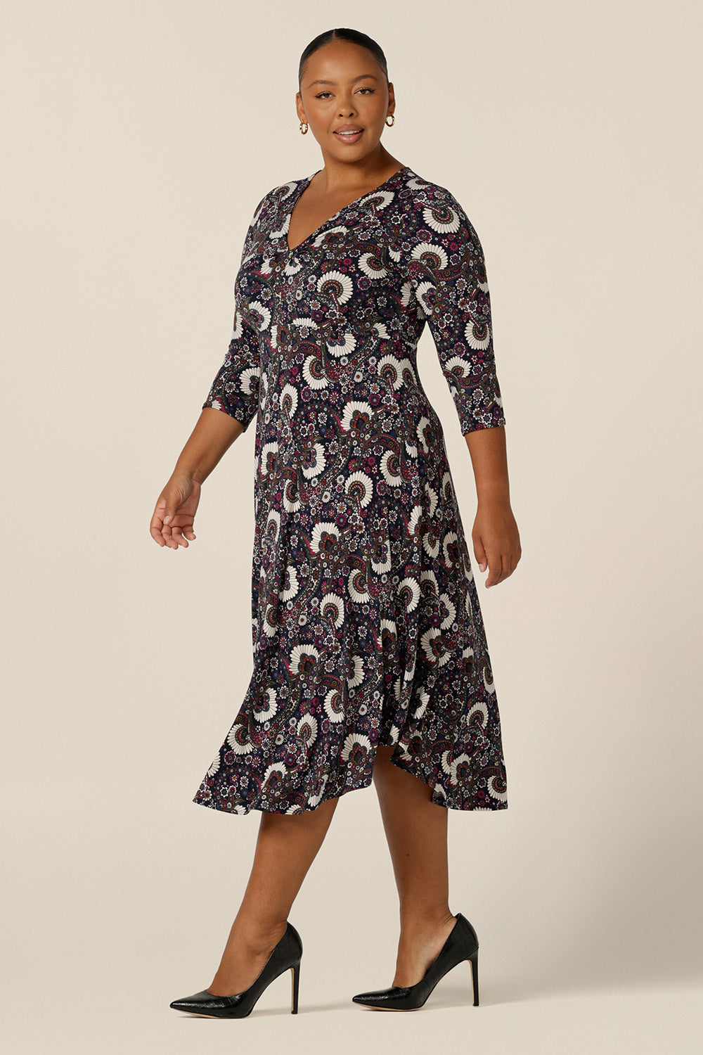 A size 18, plus size woman wears a 3/4 sleeve empire line dress with twist front bodice and dipped, calf length hem. Made in Australia by Australian and New Zealand women's clothing brand, L&F, this paisley print jersey dress is available to shop in an inclusive 8 to 24 size range.