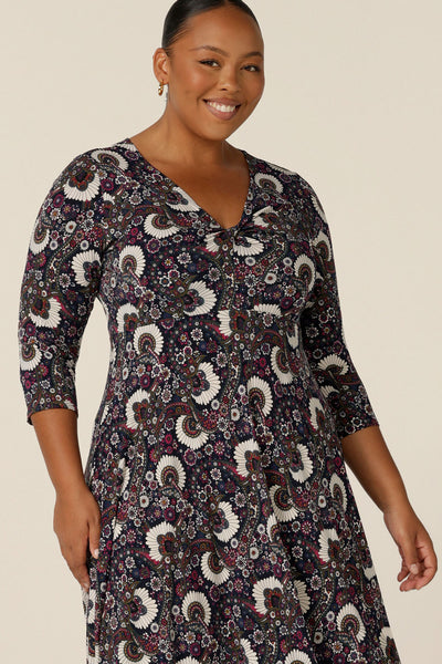 A size 18, plus size woman wears a 3/4 sleeve empire line dress with twist front bodice ad dipped, calf length hem. Made in Australia by Australian and New Zealand women's fashion label, L&F this paisley print jersey dress is available to shop in sizes 8 to 24.