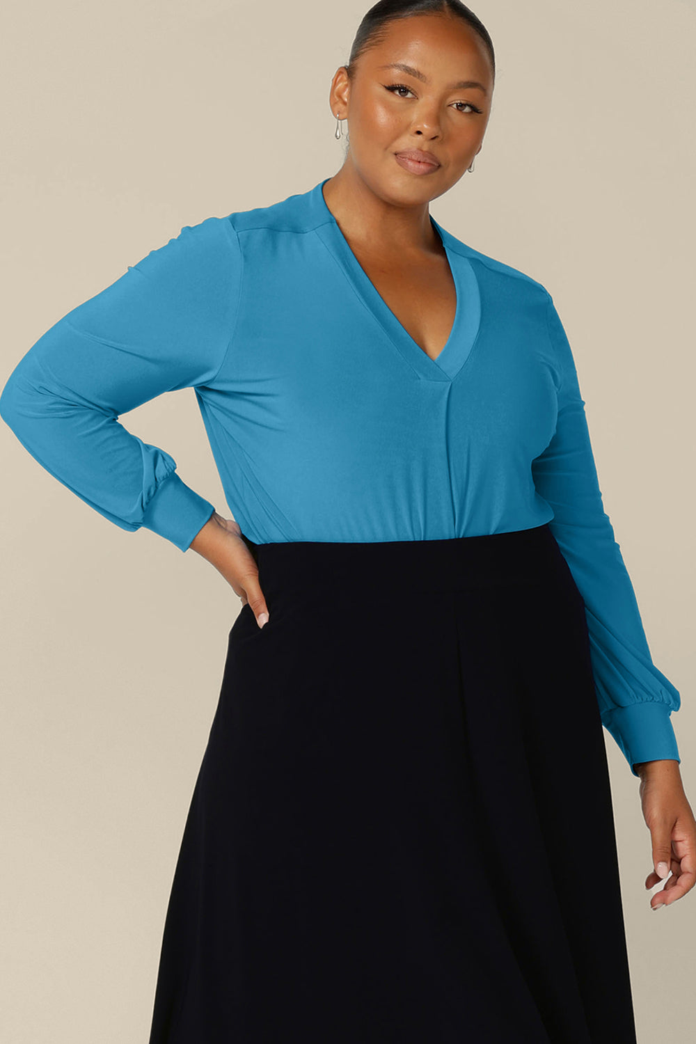 A fuller figure woman wears a V-neck top in size 18 with long bishop sleeves in Opal blue stretch jersey. Made in Australia by Australian and New Zealand women's clothing label, L&F, this workwear top is available to shop in an inclusive size range of sizes 8 to 24.