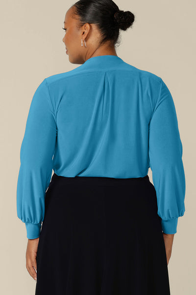 Back view of a plus size woman wearing a V-neck top in size 18 with long bishop sleeves in Opal blue stretch jersey. Made in Australia by Australian and New Zealand women's clothing label, L&F, this workwear top is available to shop in an inclusive size range of sizes 8 to 24.