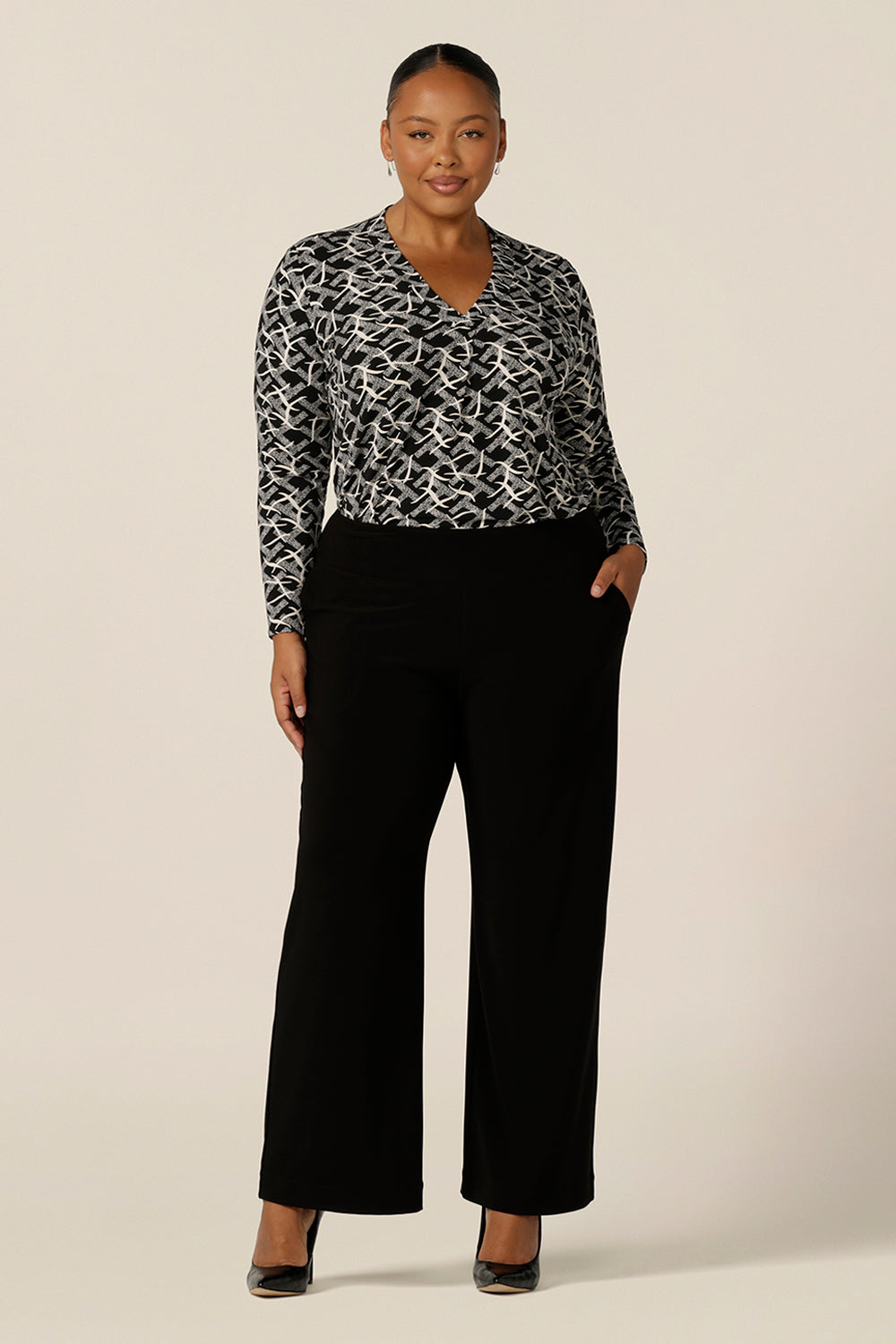 A size 18, plus size woman wears a V-neck jersey top with long sleeves and a shirttail hemline with wide leg black workwear pants. Made in Australia by women's workwear and casual wear brand, L&F, this long sleeve top features a black and white graphic print.