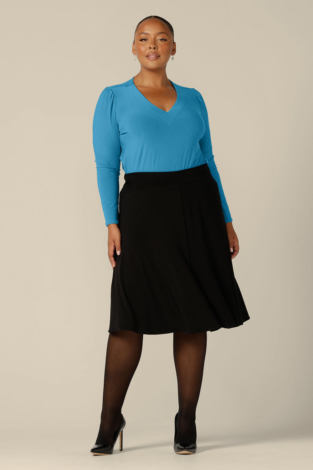 A plus size, size 18 woman wears a long sleeve, V-neck top in Opal blue jersey. Made in Australia by Australian and New Zealand women's clothing label, L&F, this top is worn with a navy blue, knee-length skirt for work. 