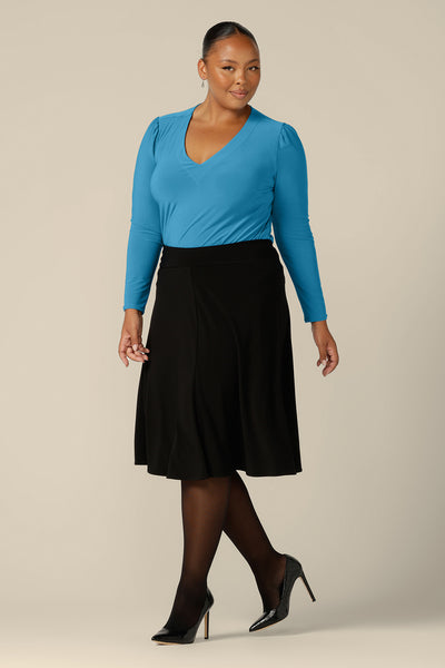 A fuller figure, size 18 woman wears a long sleeve, V-neck top in Opal blue jersey. Made in Australia by Australian and New Zealand women's clothing company, L&F, this workwear top is worn with black, knee-length skirt for work. Shop tops and skirts in inclusive sizes, 8 to 24.