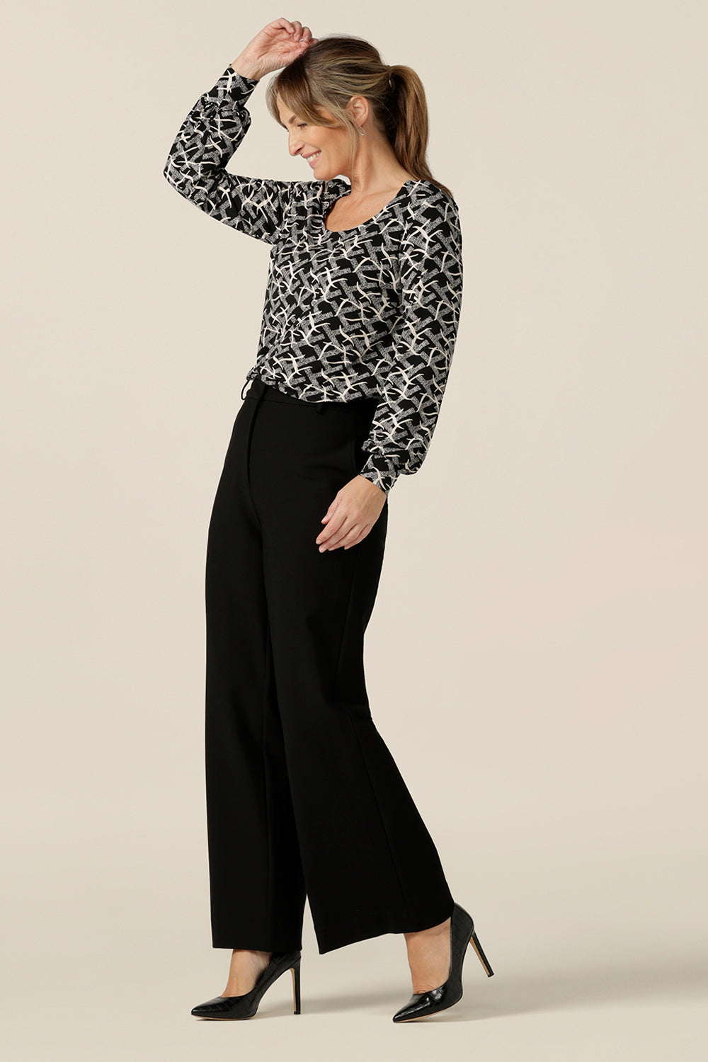 A size 10 woman wears a long sleeve top with bishop sleeve cuffs and a scoop neck in black and white print jersey with tailored, wide leg black workwear trousers. Made in Australia by Australian and New Zealand women's clothing label, L&F this women's work top is available to shop in sizes 8 to 24.