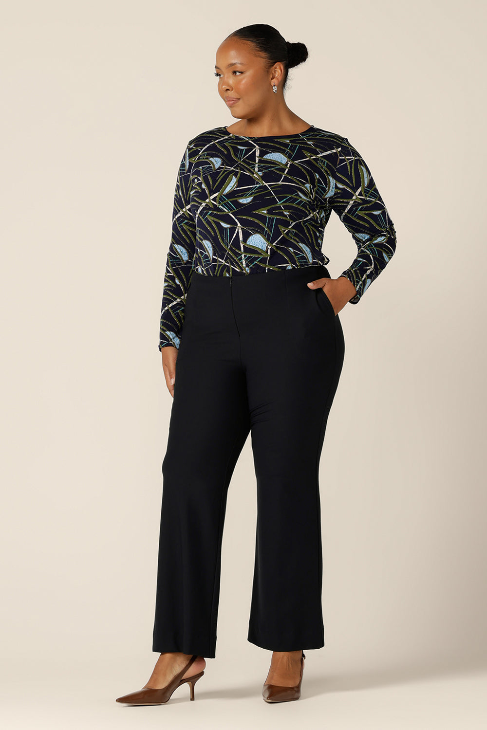 a size 18, plus size woman wears a long sleeve, boat neck top by Australian and New Zealand women's clothing brand, L&F. Featuring a blue, green and white print on a navy jersey base, this top is worn with flared leg, navy pants for a complete work or corporate wear look.