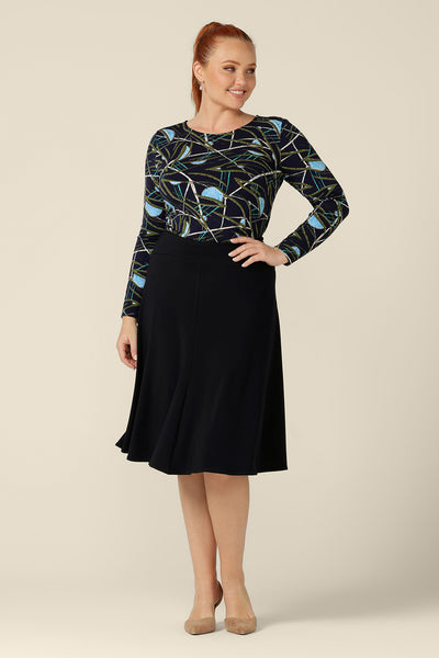a size 12, curvy woman wears a long sleeve, boat neck top by Australian and New Zealand women's clothing brand, L&F. Featuring a blue, green and white print on a navy jersey base, this top is worn with a knee-length flared skirt for a comfortable workwear outfit.
