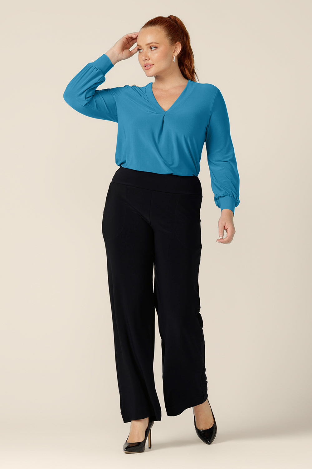 A curvy woman wears a V-neck top with long bishop sleeves in Opal blue stretch jersey. Made in Australia by Australian and New Zealand women's clothing label, L&F, this work wear top is worn with navy blue, straight leg pants for a chic work wear outfit.