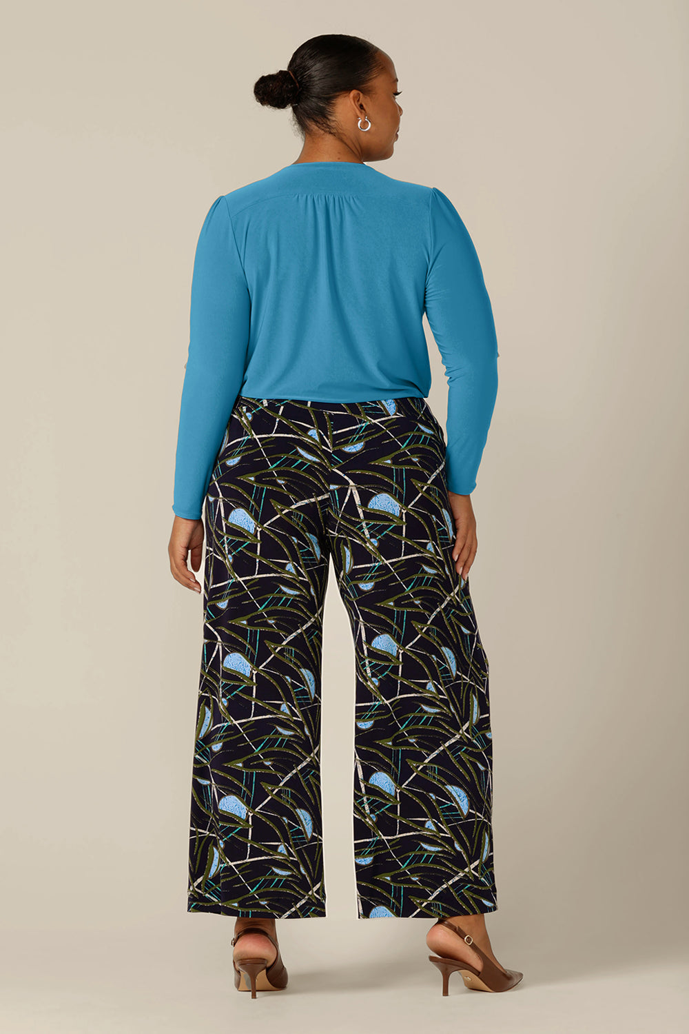 Back view of a plus size, size 18 woman wearing a long sleeve, V-neck top in Opal blue jersey with wide-leg, printed pants. Made in Australia by Australian and New Zealand women's clothing company, L&F, this top is available for shipping in sizes 8 to 24.