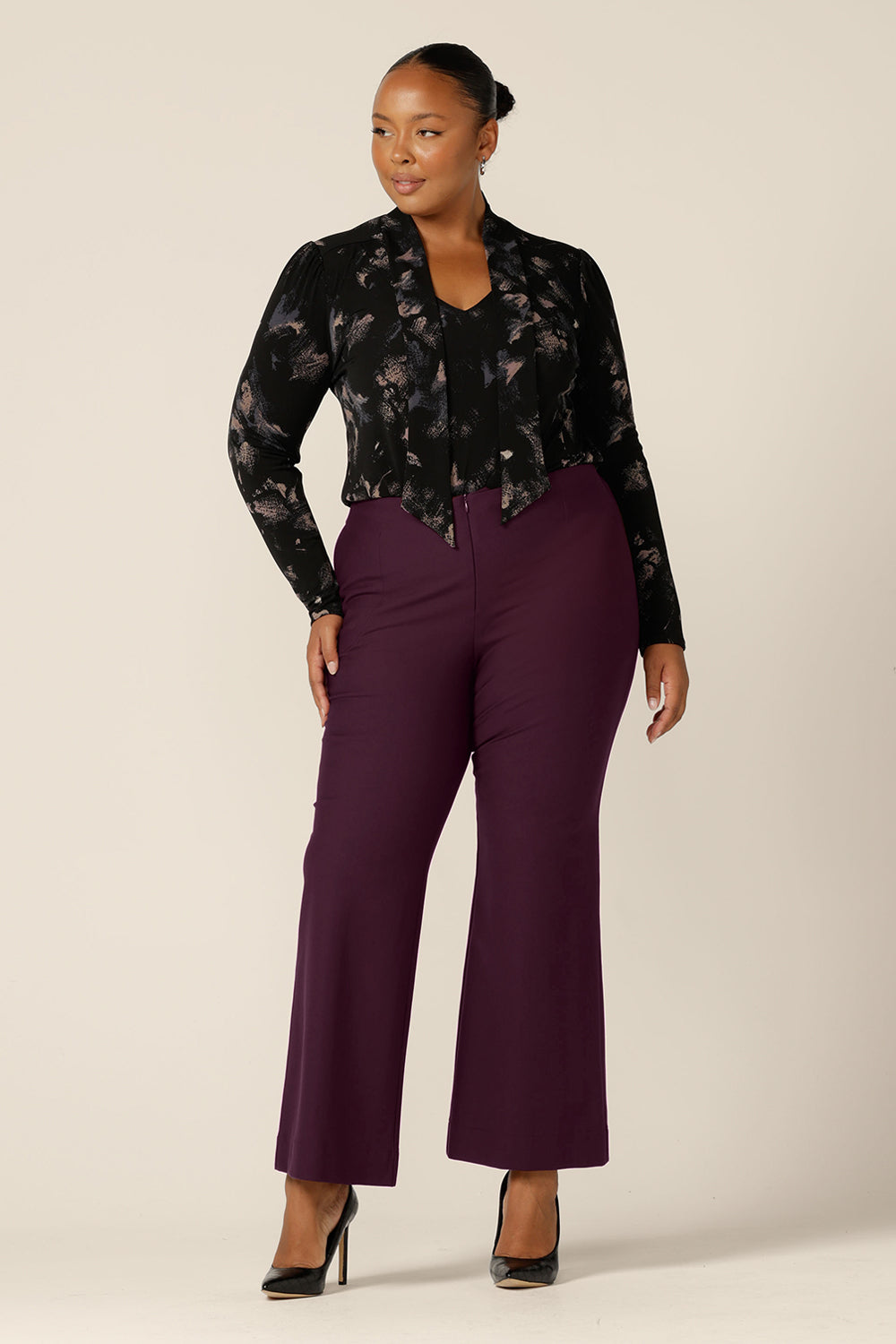 A plus size, size 18 woman wears a long sleeve, V-neck top with tie neck detail with flared leg, tailored pants in Mulberry. Both are by Australian and New Zealand women's clothing label, L&F and available to shop in sizes 8 to 24.