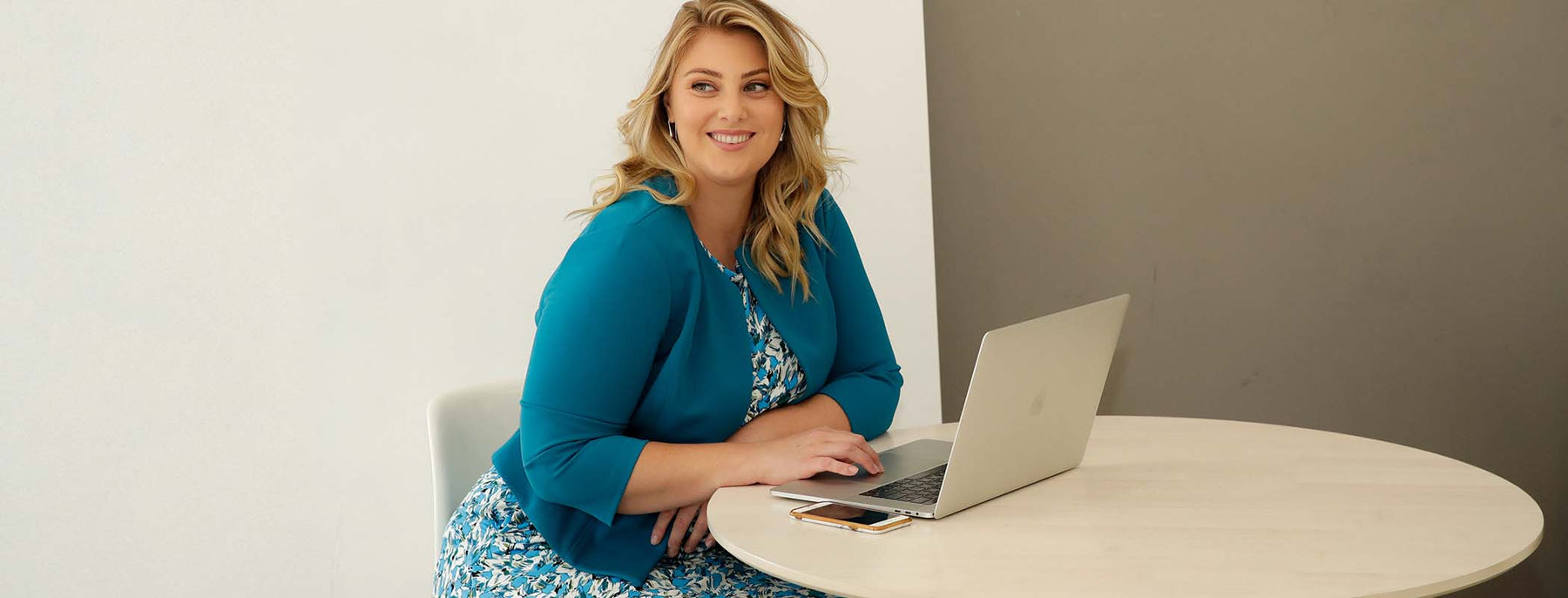 Plus size woman sitting at office desk wearing a printed jersey work dress with blue tailored jacket to show made in Australia corporate looks