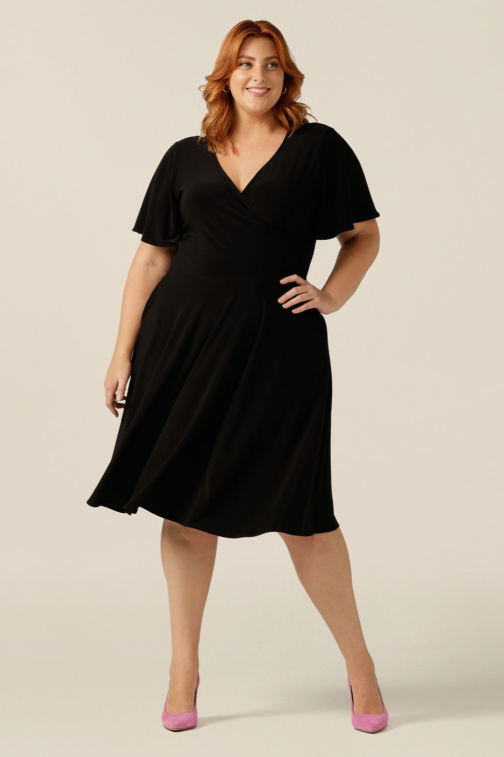 size 18, plus size woman wearing a reversible fixed wrap dress in black jersey. Worn reversed, she wears it as a boat-neck little black dress. Featuring short flutter sleeves, a knee length skirt and pockets, this dress can be worn for work wear or going-out dress style. Made in Australia by women's clothing label, Leina and Fleur