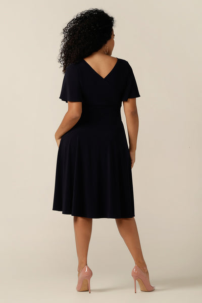 size 12, curvy woman wearing a reversible fixed wrap dress in navy jersey. The dress features flutter sleeves, knee length skirt and pockets. the reversible style of the dress allows her to be worn with a wrap front (as shown) or as a boat-neck dress. Designed as a work wear or evening dress, it is made in Australia for petite to plus sizes by women's clothing label, Leina and Fleur