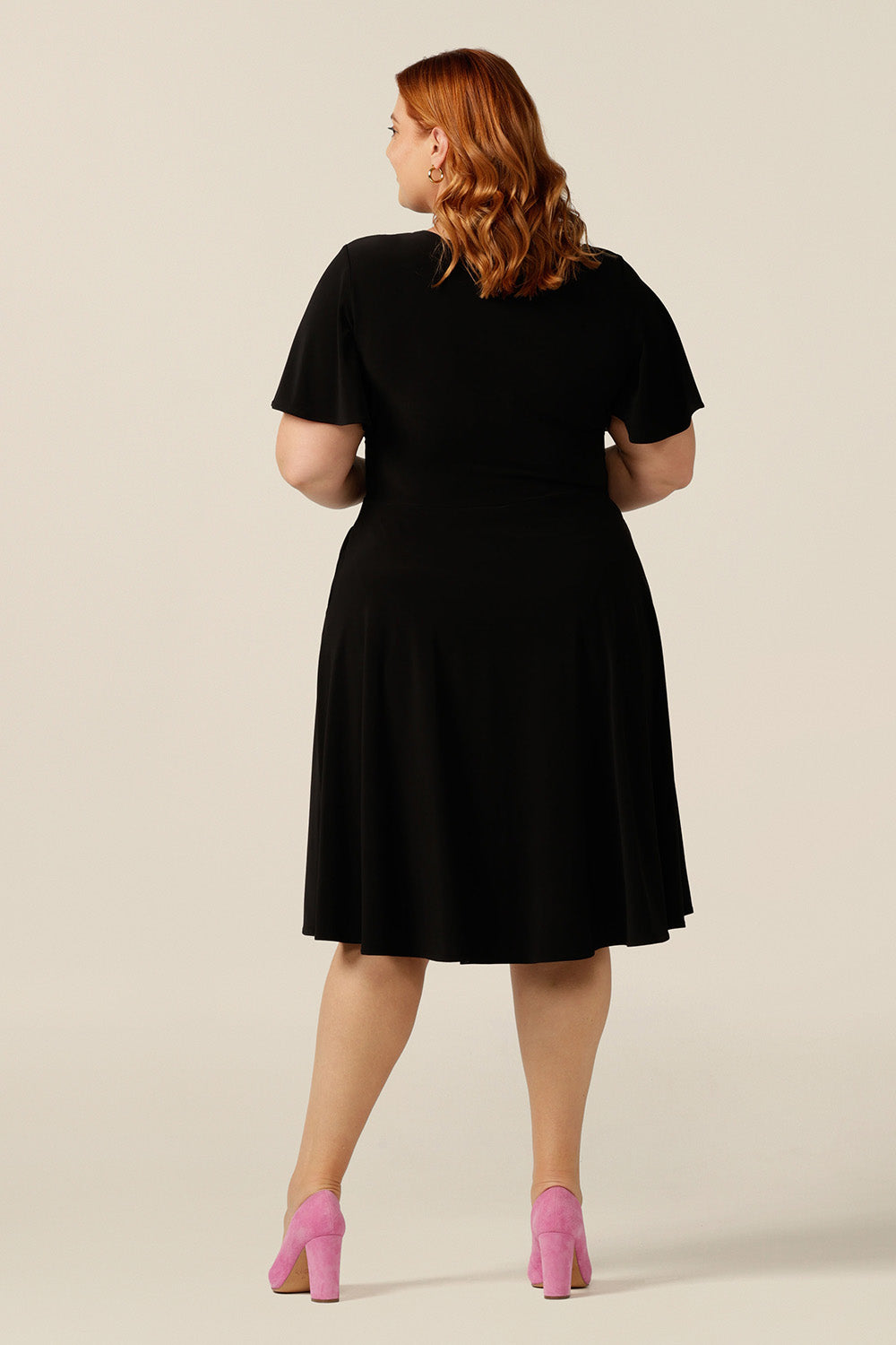 size 18, plus size woman wearing a reversible fixed wrap dress in black jersey. Worn with the wrap front forwards, she styles as elegant jersey little black dress. Featuring short flutter sleeves, a knee length skirt and pockets, this dress can be worn for work wear or going-out dress style. Made in Australia by women's clothing label, Leina and Fleur