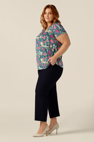 Size 18 curvy woman wearing a semi-fitted V-neck top with short sleeves. The top is shown in floral print slinky jersey for a feminine work wear top. Worn with tailored navy pants, the image shows a successful pairing of smart-casual work wear separates.. Both the top and trousers are made in Australia by petite-to-plus-size clothing experts, Leina and Fleur