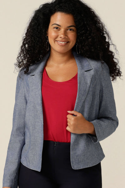 A size 12 curvy woman wears a softly tailored jacket in cotton-linen blenc Chambray fabric. the jacket is open-fronted and has long sleeves. Under the jacket, the woman wears a red bamboo jersey top with round neck, and black trousers. The jacket is styled as a smart-casual cover-up for weekend and going out fashion.