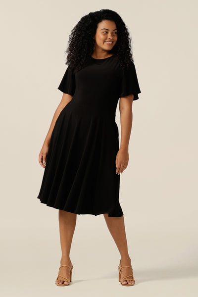 size 12, curvy woman wearing a reversible fixed wrap dress in black jersey. Worn reversed, as a boat neck dress, she styles as elegant jersey little black dress. Featuring short flutter sleeves, a knee length skirt and pockets, this dress can be worn for work wear or going-out dress style. Made in Australia by women's clothing label, Leina and Fleur