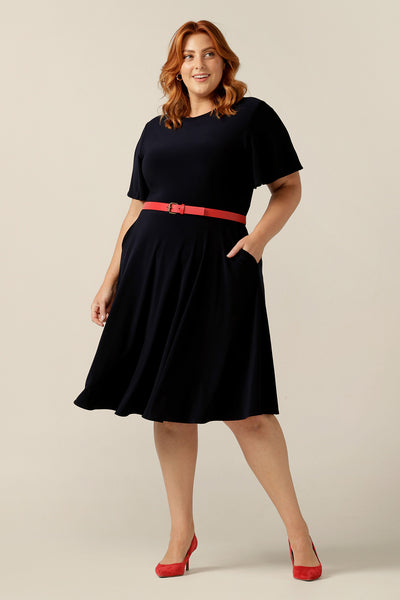 size 18, curvy woman wearing a reversible fixed wrap dress in navy jersey. The dress features flutter sleeves, knee length skirt and pockets. the reversible style of the dress allows her to be worn with a wrap front (as shown) or as a boat-neck dress. Designed as a work wear or evening dress, it is made in Australia for petite to plus sizes by women's clothing label, Leina and Fleur