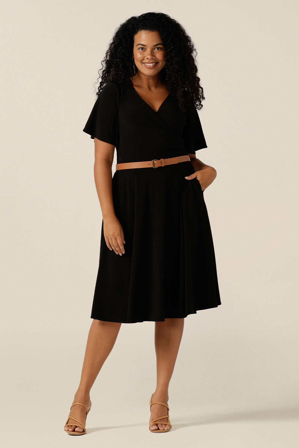 size 12, curvy woman wearing a reversible fixed wrap dress in black jersey. Worn with the wrap front forwards, she styles as an elegant jersey little black dress. Featuring short flutter sleeves, a knee length skirt and pockets, this dress can be worn for work wear or going-out dress style. Made in Australia by women's clothing label, Leina and Fleur