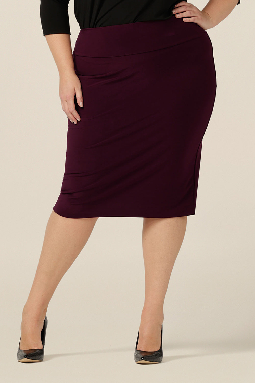 A size 18, plus size woman wears a knee-length tube skirt in Mulberry stretch jersey. A workwear pencil skirt, the stretch fabric makes for a comfortable skirt for all-day corporate wear.