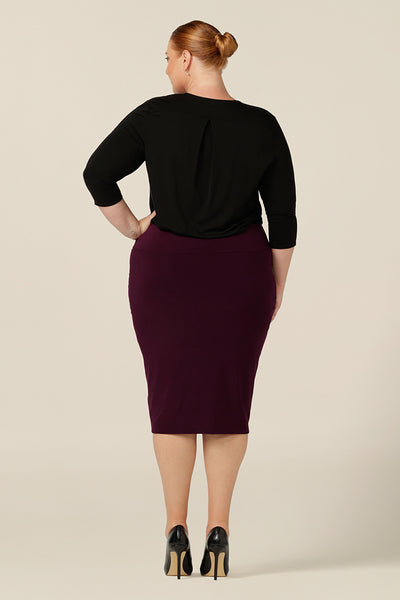 Back view of a size 18, fuller figure woman wearing a tube skirt in Mulberry stretch jersey. A knee-length pencil skirt styled with a long sleeve black top, the stretch fabric makes for a comfortable skirt for workwear.