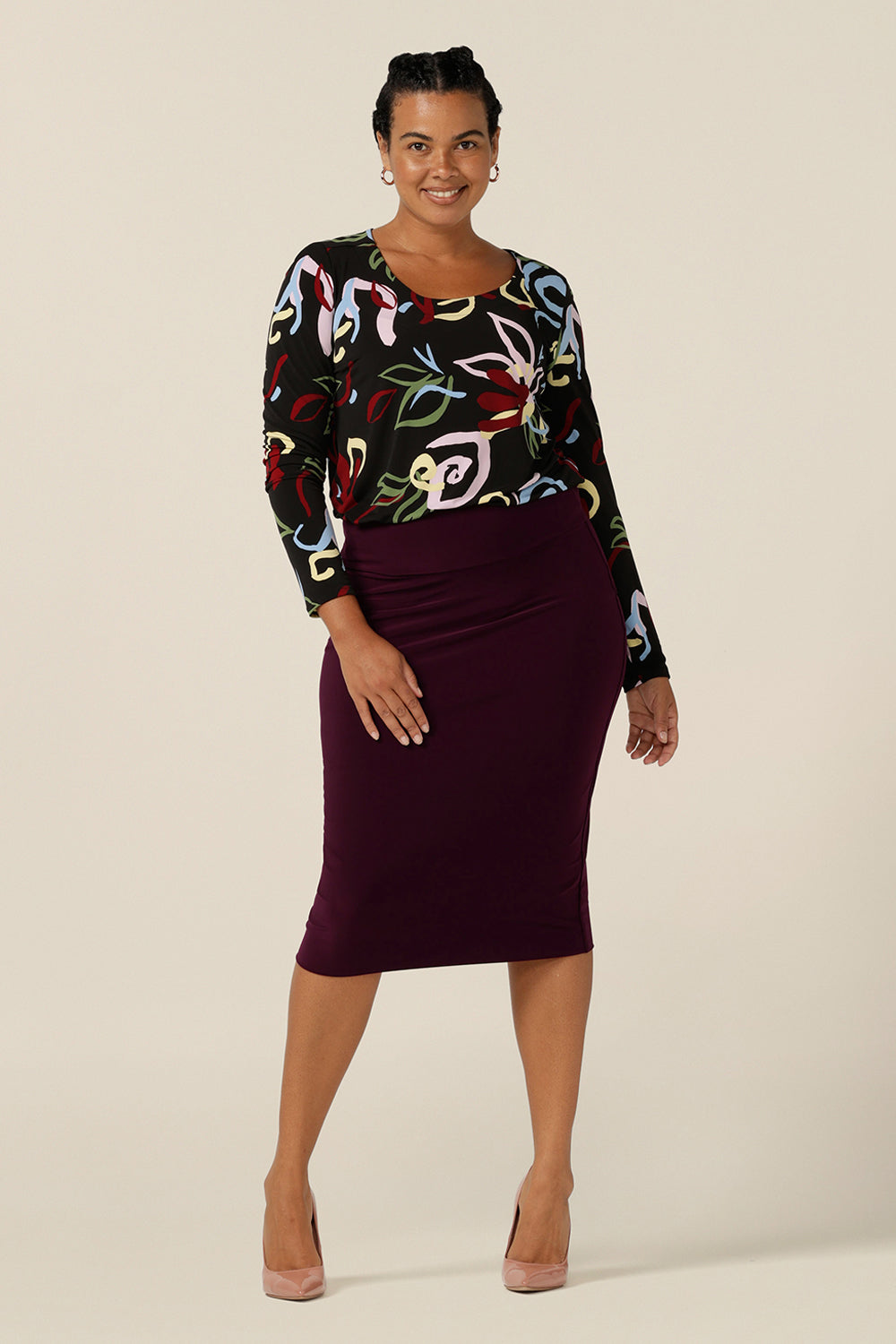 Styled for work, a round neck jersey top with abstract print and full length sleeves is worn tucked in to a workwear tube skirt in Mulberry. Shown worn by a size 12 woman, shop this work top in an inclusive size 8 to size 24 range. 