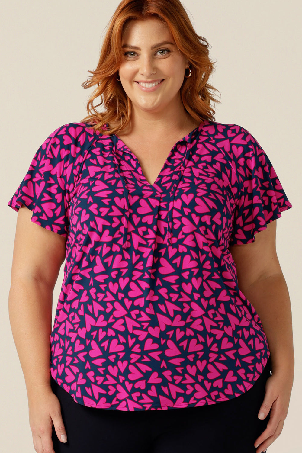 A plus size, size 18 woman wears a casual jersey top. The top has short raglan sleeves, a V-neckline and is printed with pink hearts for a Valentines Day fashion look. 