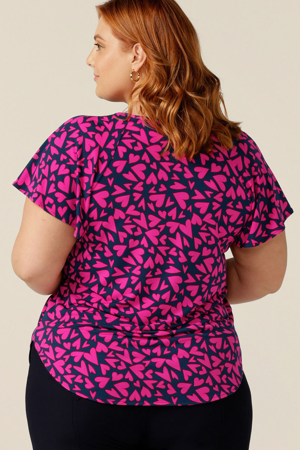 A plus size, size 18 woman wears a casual jersey top. The top has short raglan sleeves, a V-neckline and is printed with pink hearts for a Valentines Day fashion look. 